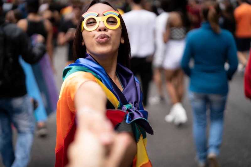 A young feminine-presenting person in a rainbow cape and yellow goggles holds a hand out in invitation to the viewer. Crowds in the background appear to be celebrating.