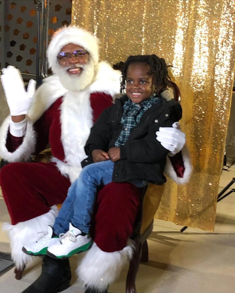 A young child in neatly plaited hair sits on the lap of a Santa Claus character in a red suit with whit trim, a red and white hat and white gloves. Both the boy and the actor are Black.