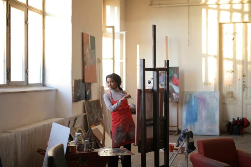 Black female artist sitting at an easel painting.