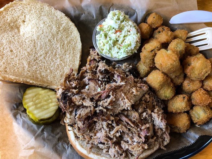 A plateful with a chopped pork BBQ sandwich with cole slaw, pickles, and fried okra.