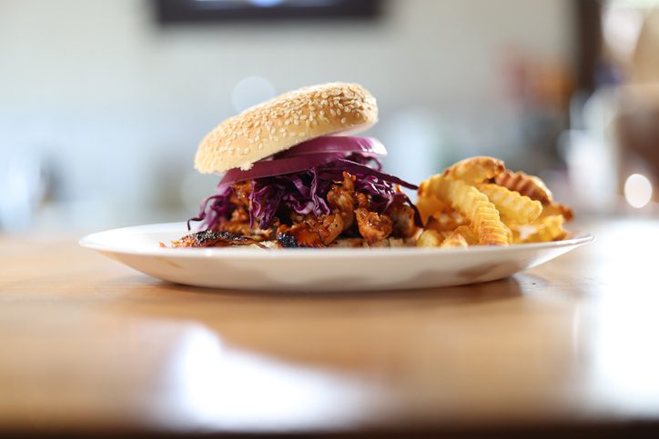 chargrilled chicken BBQ sandwich garnished with onions and coleslaw is served next to some salty french fries presented on a white plate, as found in the restaurants with the best BBQ in Winston-Salem