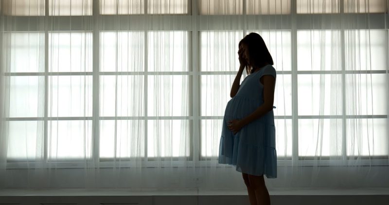 Photo of a pregnant rubbing her stomach and holding her head in a low-light room.